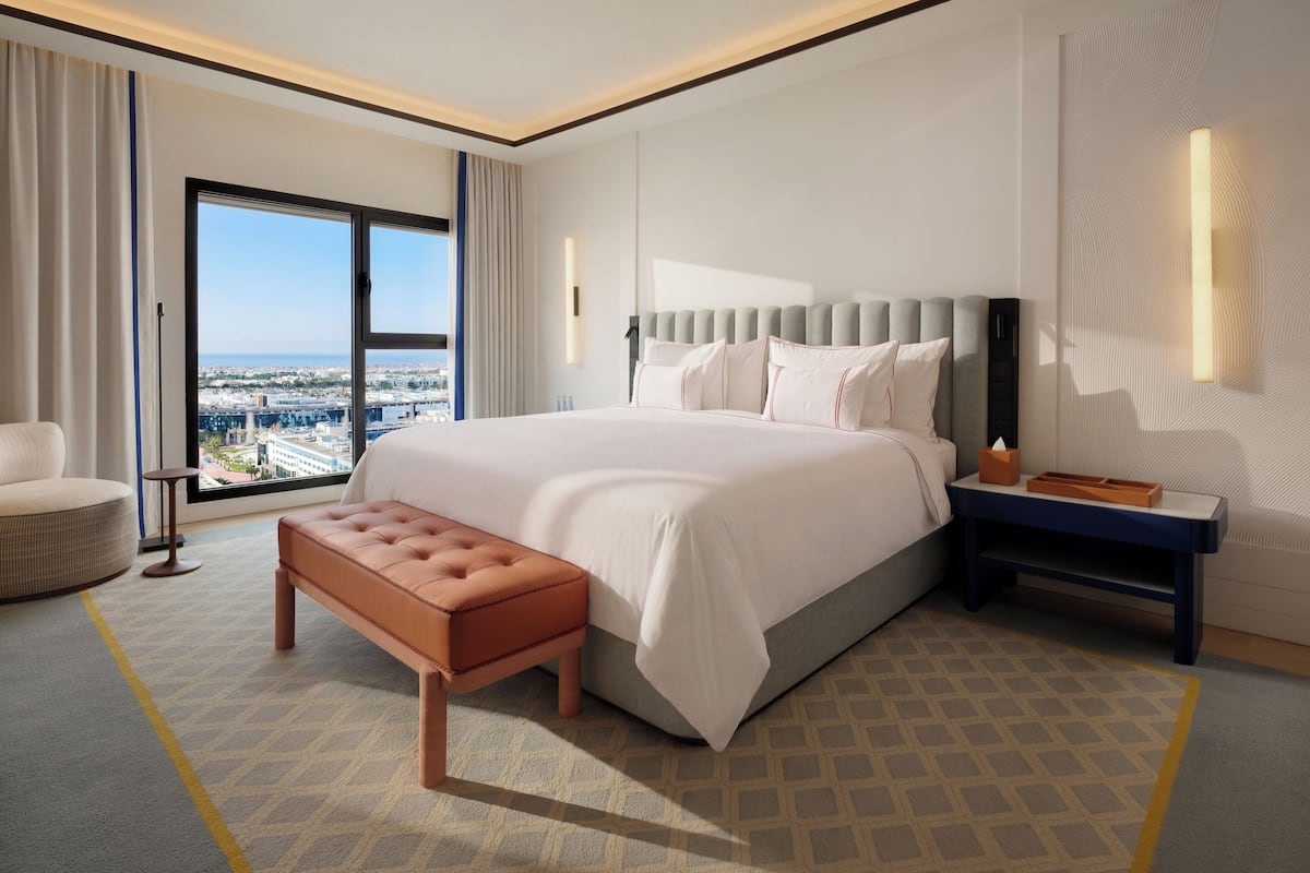 Una habitación de hotel moderna con una cama grande, un banco, una mesita de noche y vistas a la ciudad a través de un ventanal amplio.