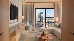 A modern hotel room with a large window, a flat-screen TV, a sofa, a coffee table with fruit, and a view of the cityscape. - The View Rabat (Rabat)