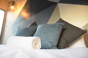 A neatly made bed with pillows and a rolled towel, a geometric-patterned wall, and a hanging light fixture.