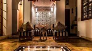 Un interior tradicional marroquí con una pared con motivos, un sofá central con cojines, dos sillones y una mesita. En primer plano, una fuente decorativa con velas.