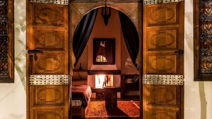 Una puerta de madera con intrincados grabados y refuerzos metálicos, que da paso a un cuarto cálidamente iluminado con chimenea, cama y una mesita.