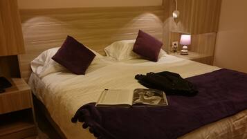 A hotel room with a wooden headboard, a bed with white and purple bedding, a purple throw blanket, a black bag, and a magazine on the bed.