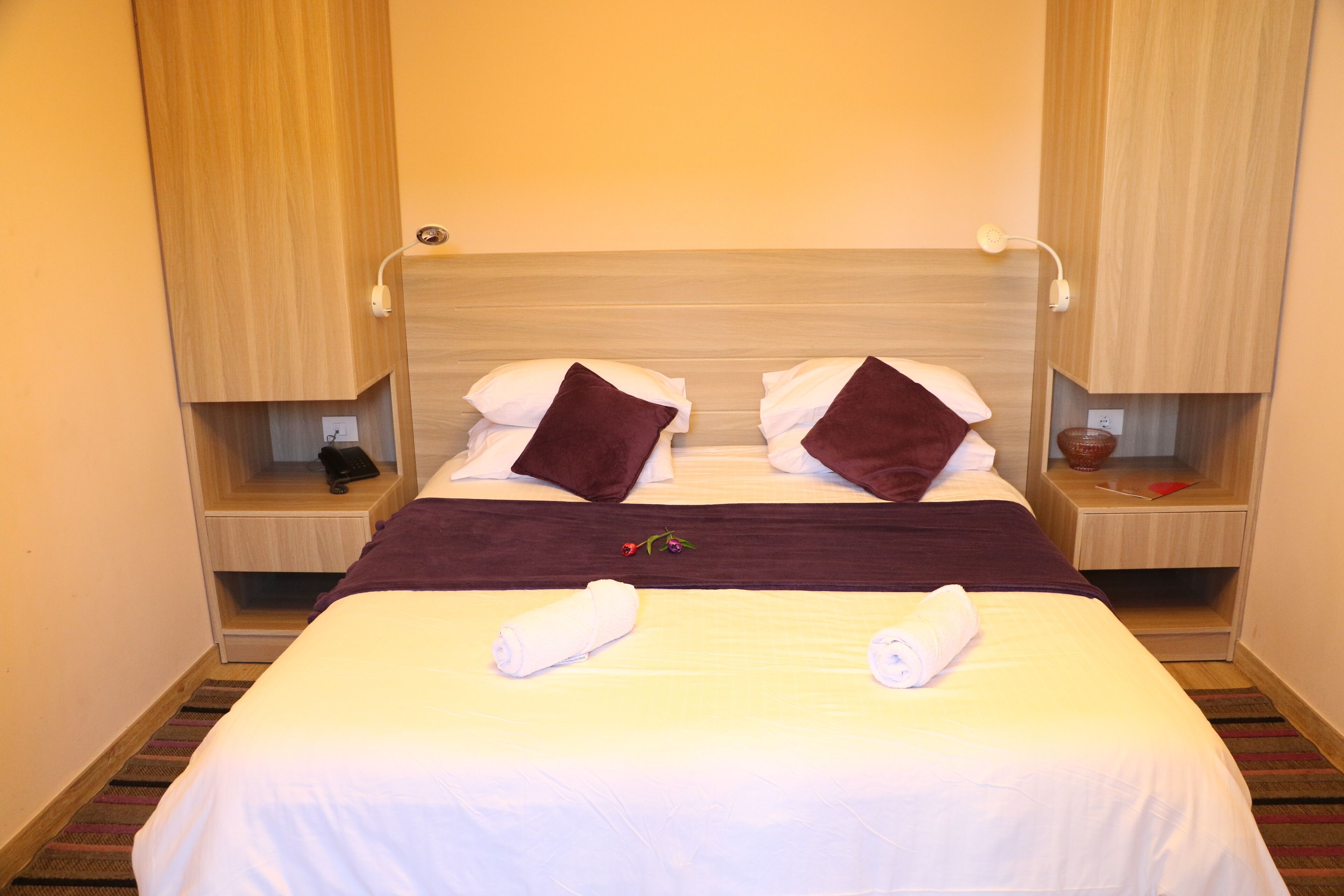 A neatly made bed with two pillows, a blanket, and two towels. There are bedside tables with a lamp and a phone.