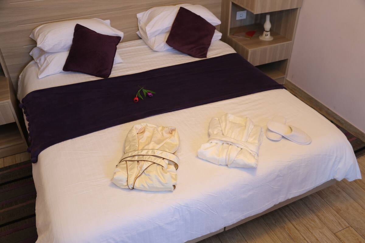 A neatly made bed with white linens, a dark-colored blanket, and two towels folded on the bed.