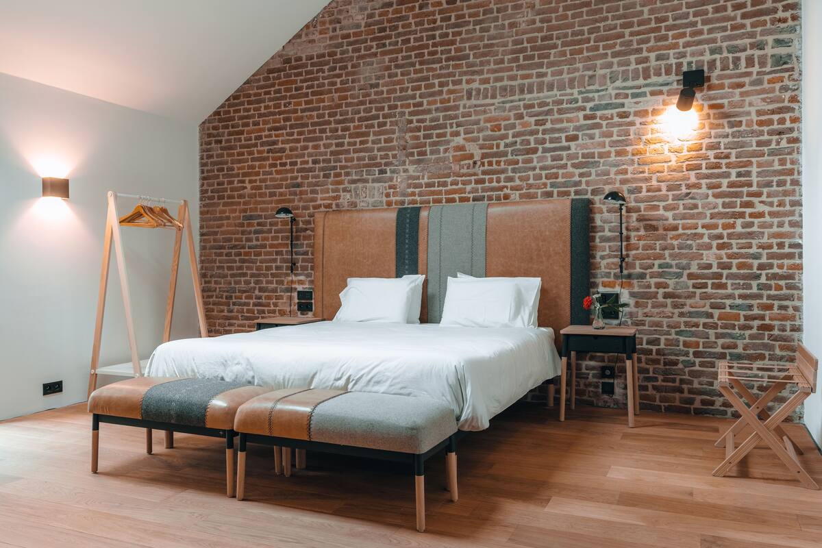 A bedroom with a brick wall, a bed with a wooden headboard, a bench, and a small table with a lamp.