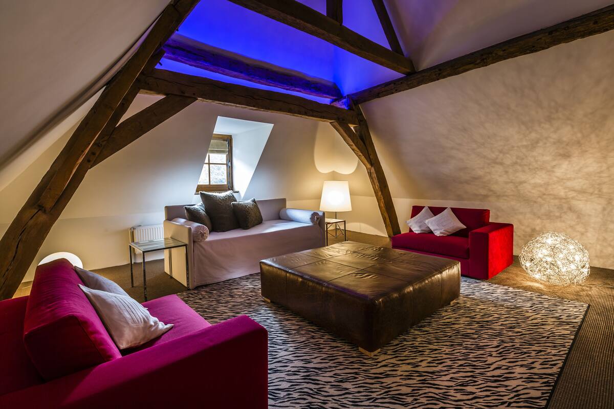 A cozy attic bedroom with a sloped ceiling, a red sofa, a black leather ottoman, and a patterned rug.