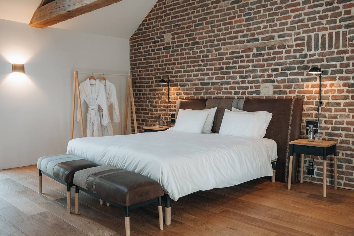 A bedroom with a brick wall, a bed with white bedding, a bench, and bedside tables with lamps.