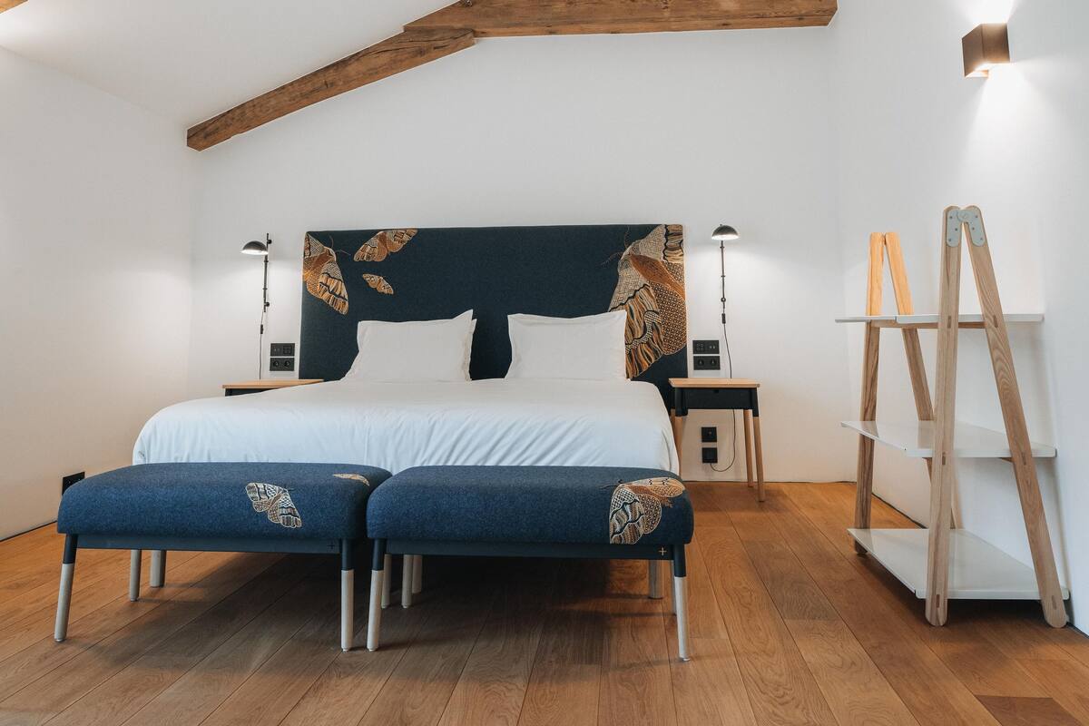 A bedroom with a wooden ceiling, a bed with a dark headboard, bedside tables, and a wooden ladder shelf.