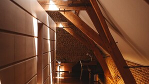 A narrow, wooden corridor with sloped ceilings and exposed brick walls, leading to a room with a brick wall and a window. - Indrani Lodge (Genappe)