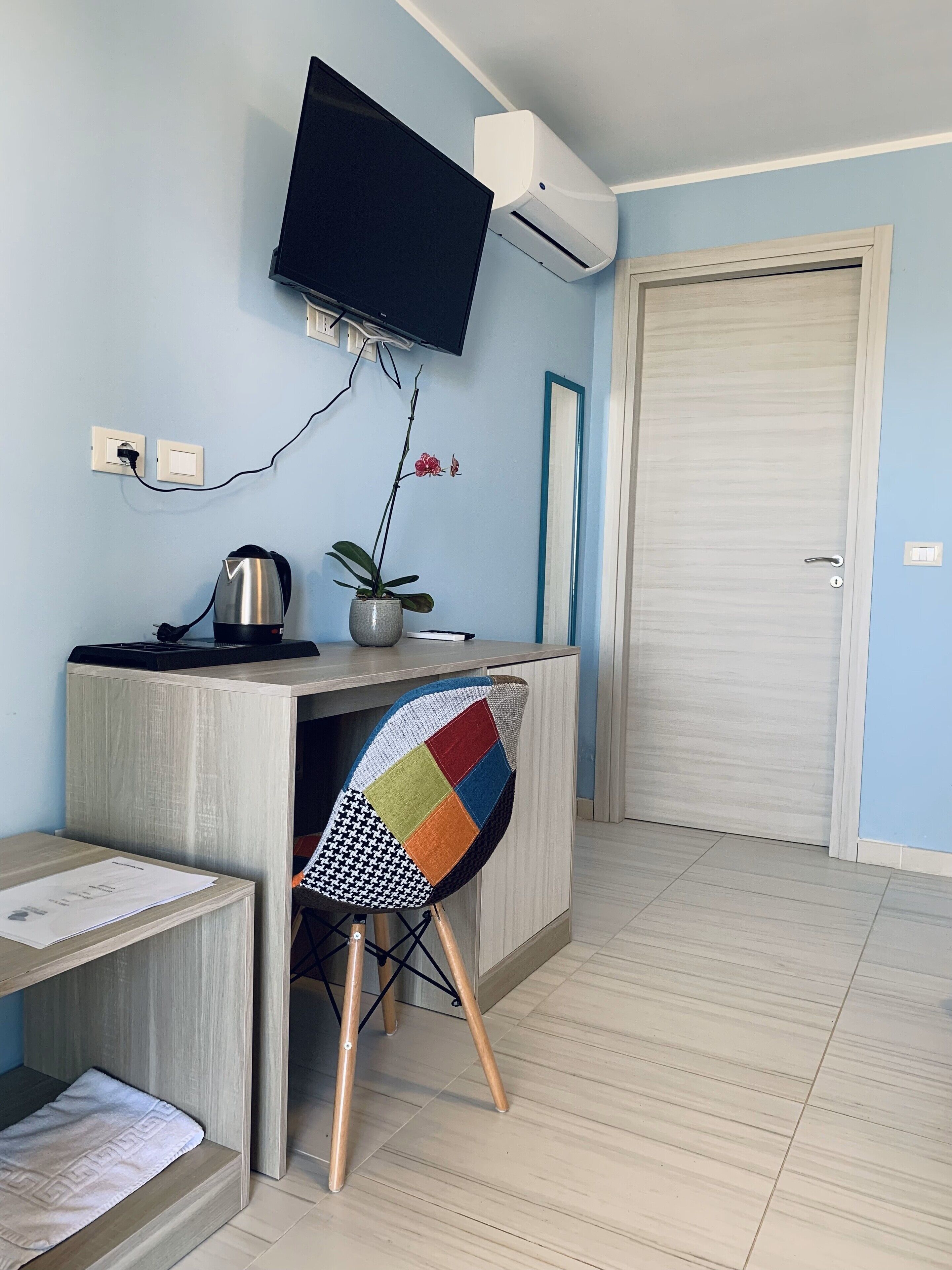 A room with a wooden desk, a flat-screen TV mounted on the wall, a potted plant, and a colorful chair.