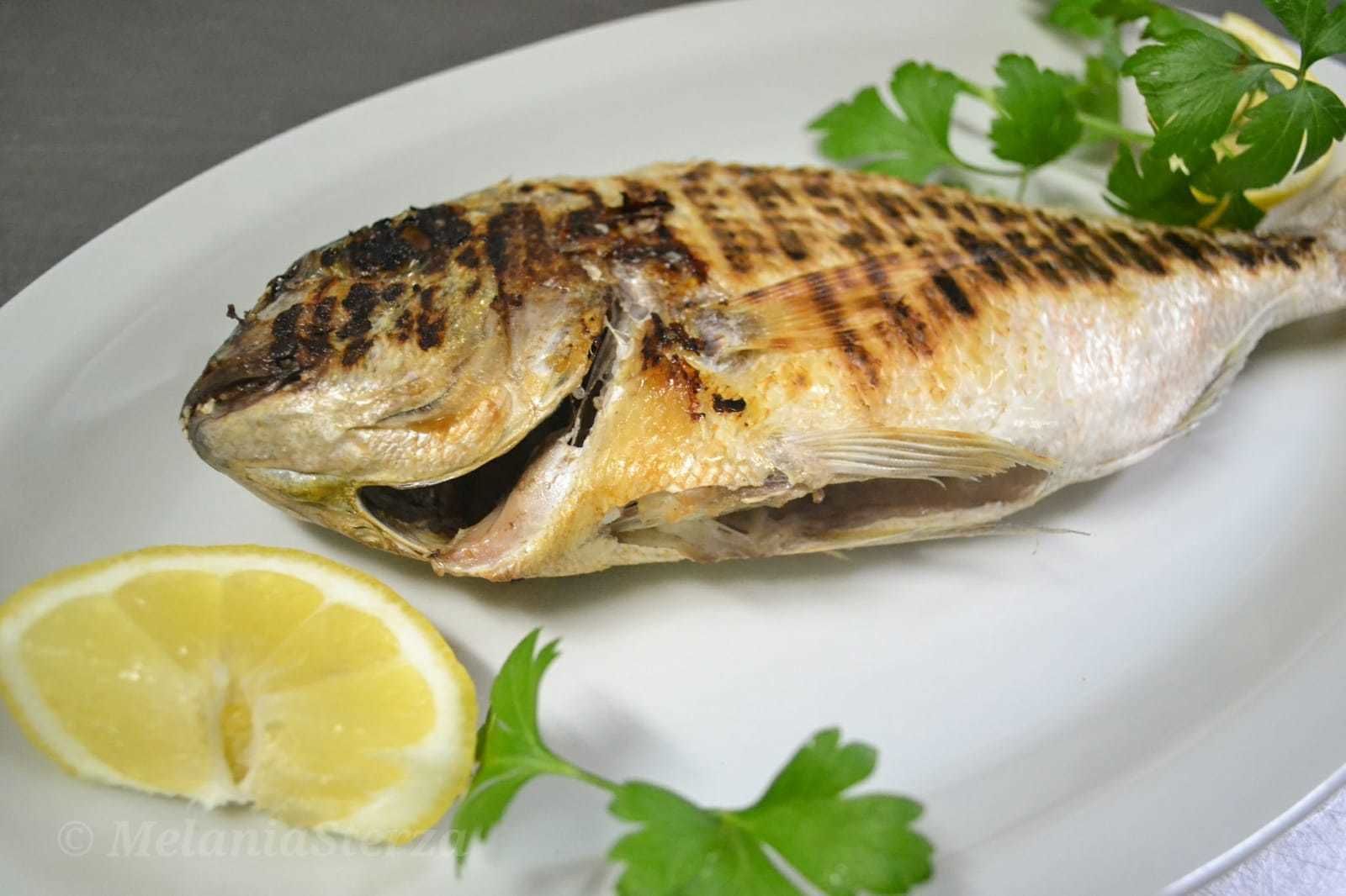 A grilled fish on a plate with a lemon wedge and parsley.