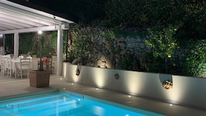 A swimming pool with a stone wall and plants, a covered seating area, and a stone building in the background. - Hotel Mercurio (Joppolo)