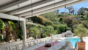 An outdoor dining area with a long table set for a meal, surrounded by chairs, with a pool and greenery in the background. - Hotel Mercurio (Joppolo)