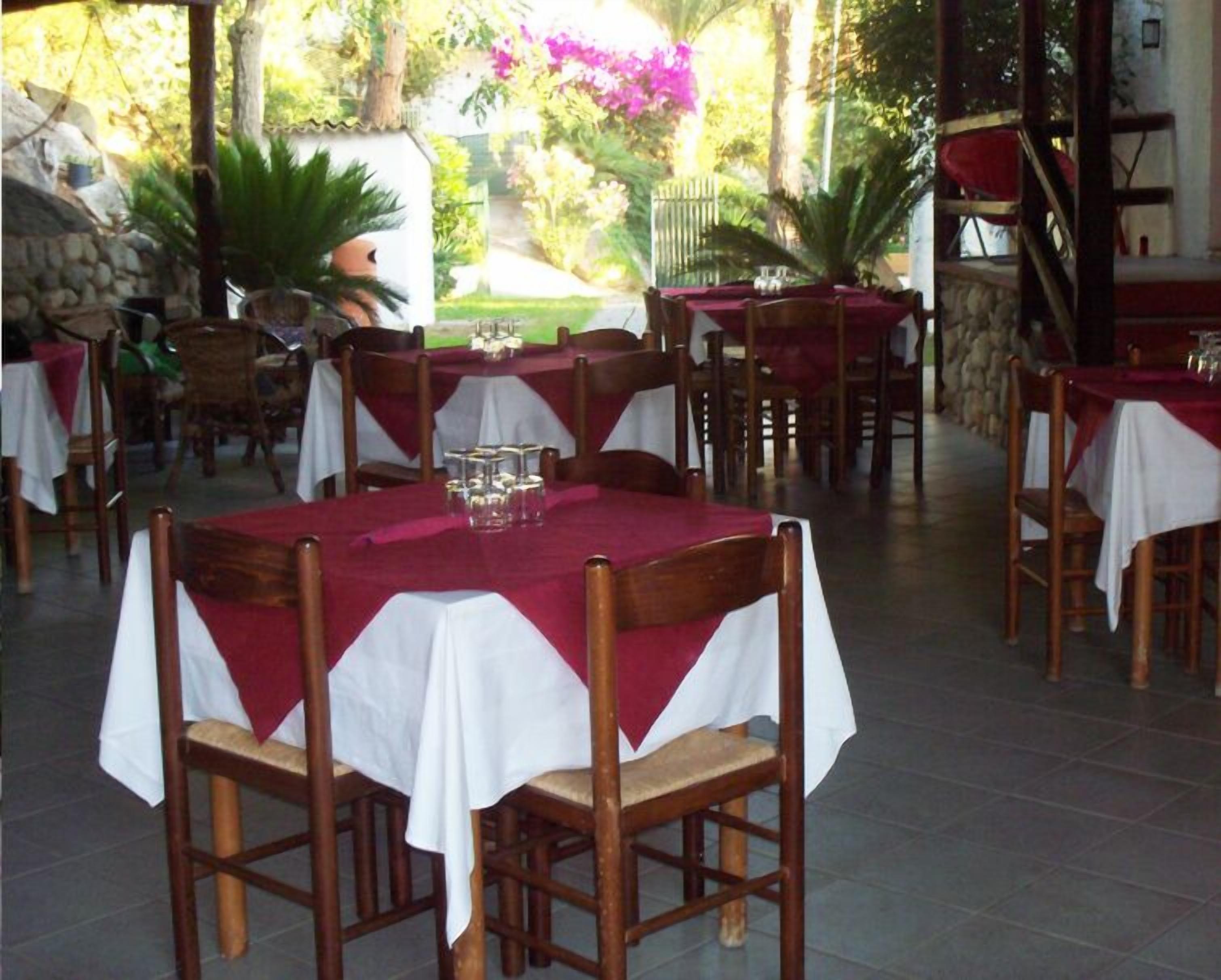 A restaurant with outdoor seating, tables covered with white tablecloths and red napkins, surrounded by greenery and a stone wall.