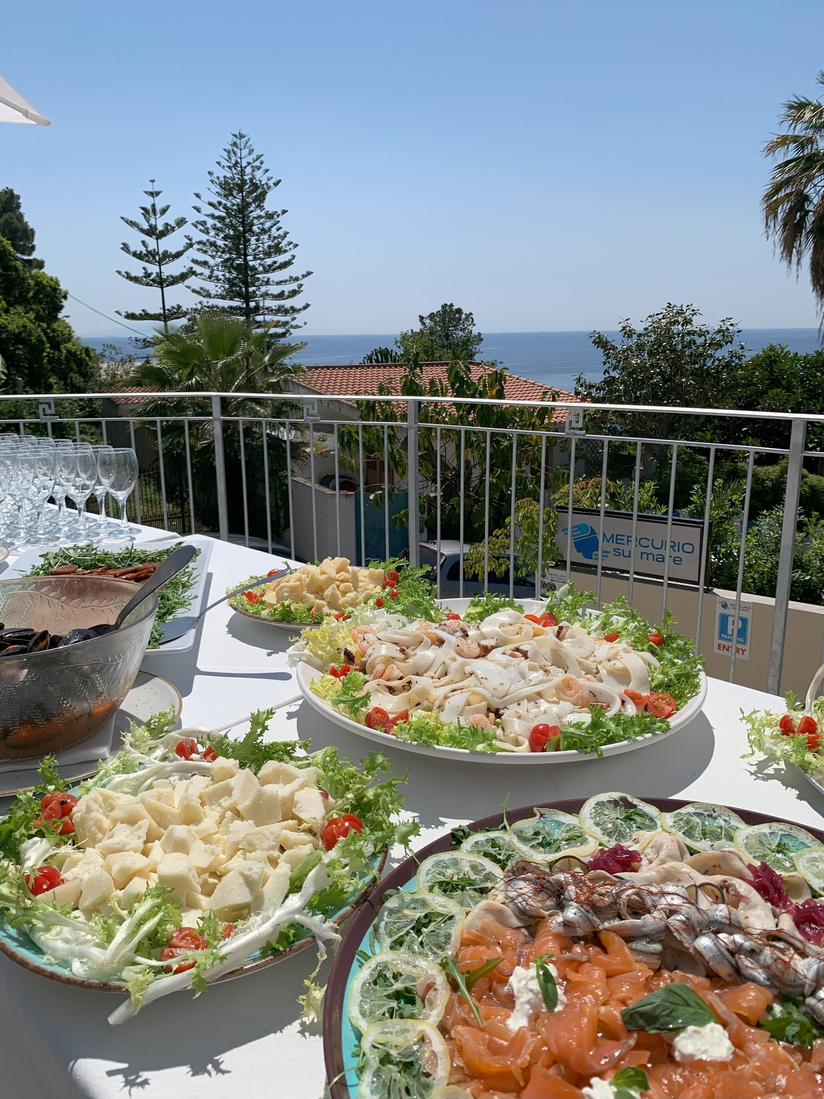 A terrace with various dishes, including seafood salads and grilled items, overlooking a scenic view with trees and a building in the background.