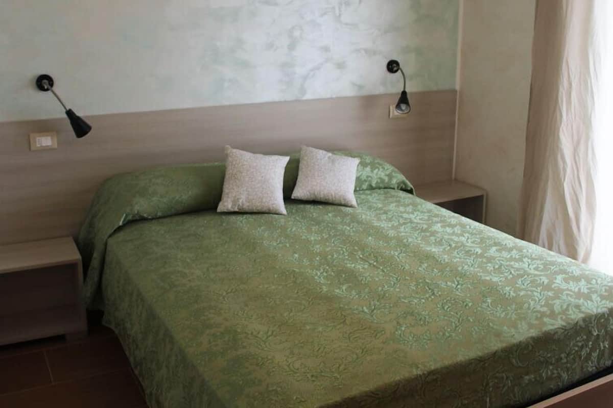 A bedroom with a green patterned bedspread, two pillows, a nightstand, and a wall-mounted lamp.