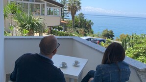 A balcony with two white plastic chairs, a small white table with two white cups, and a view of the sea and greenery. - Hotel Mercurio (Joppolo)