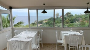 A dining area with tables set for two, large windows offering a view of the sea and greenery. - Hotel Mercurio (Joppolo)