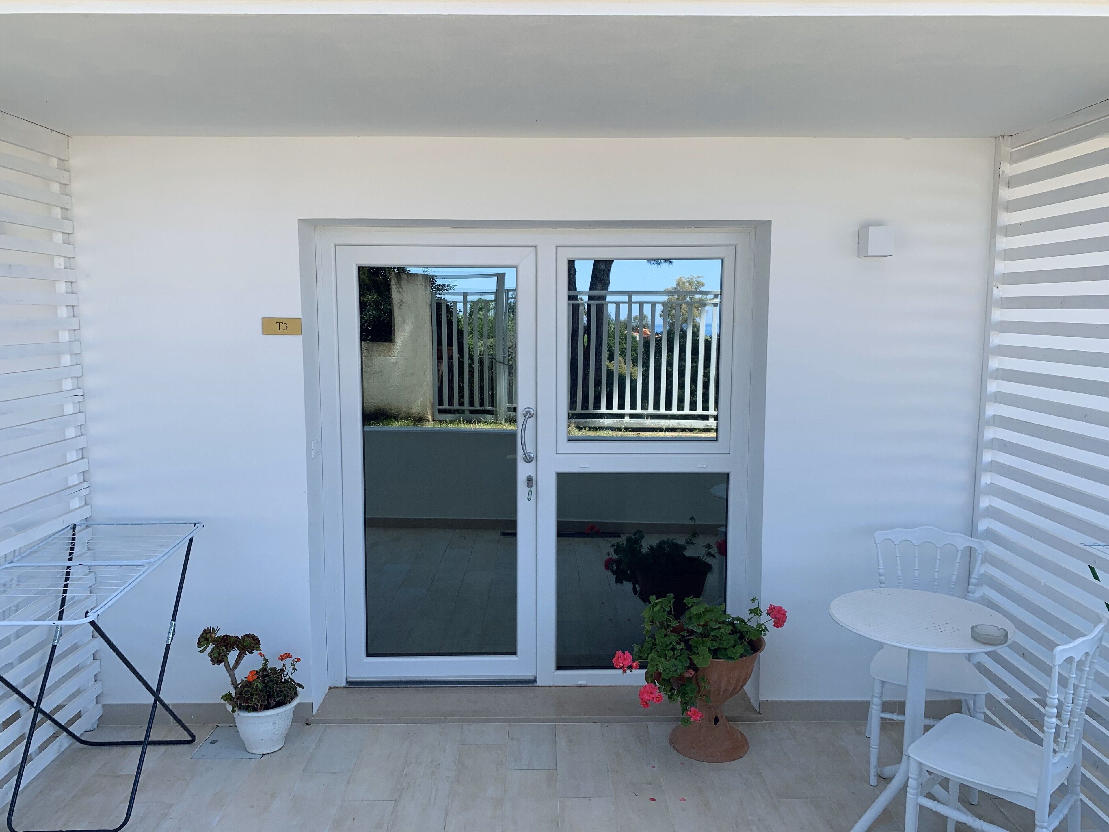 A balcony with a white door, a potted plant, a drying rack, and a white table and chair.