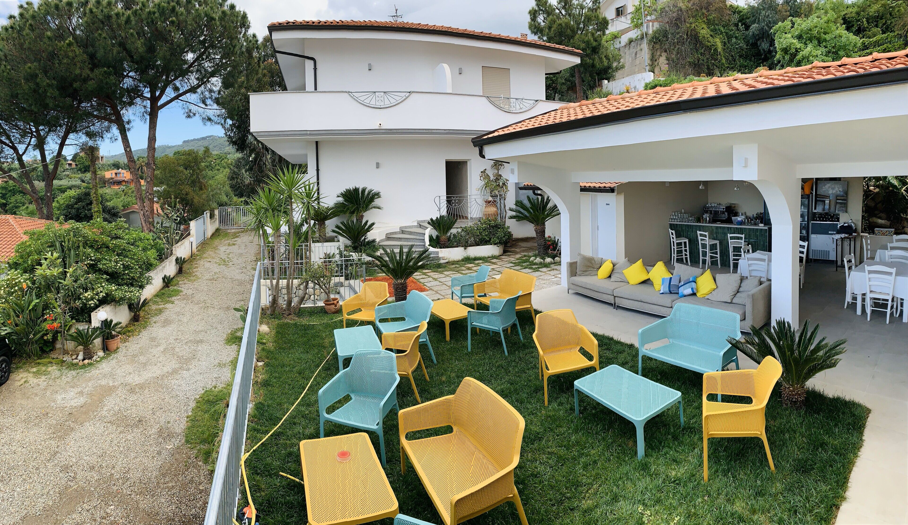 A modern outdoor seating area with a mix of yellow and blue chairs and tables, a white building with a balcony, and a kitchen area visible through an archway.