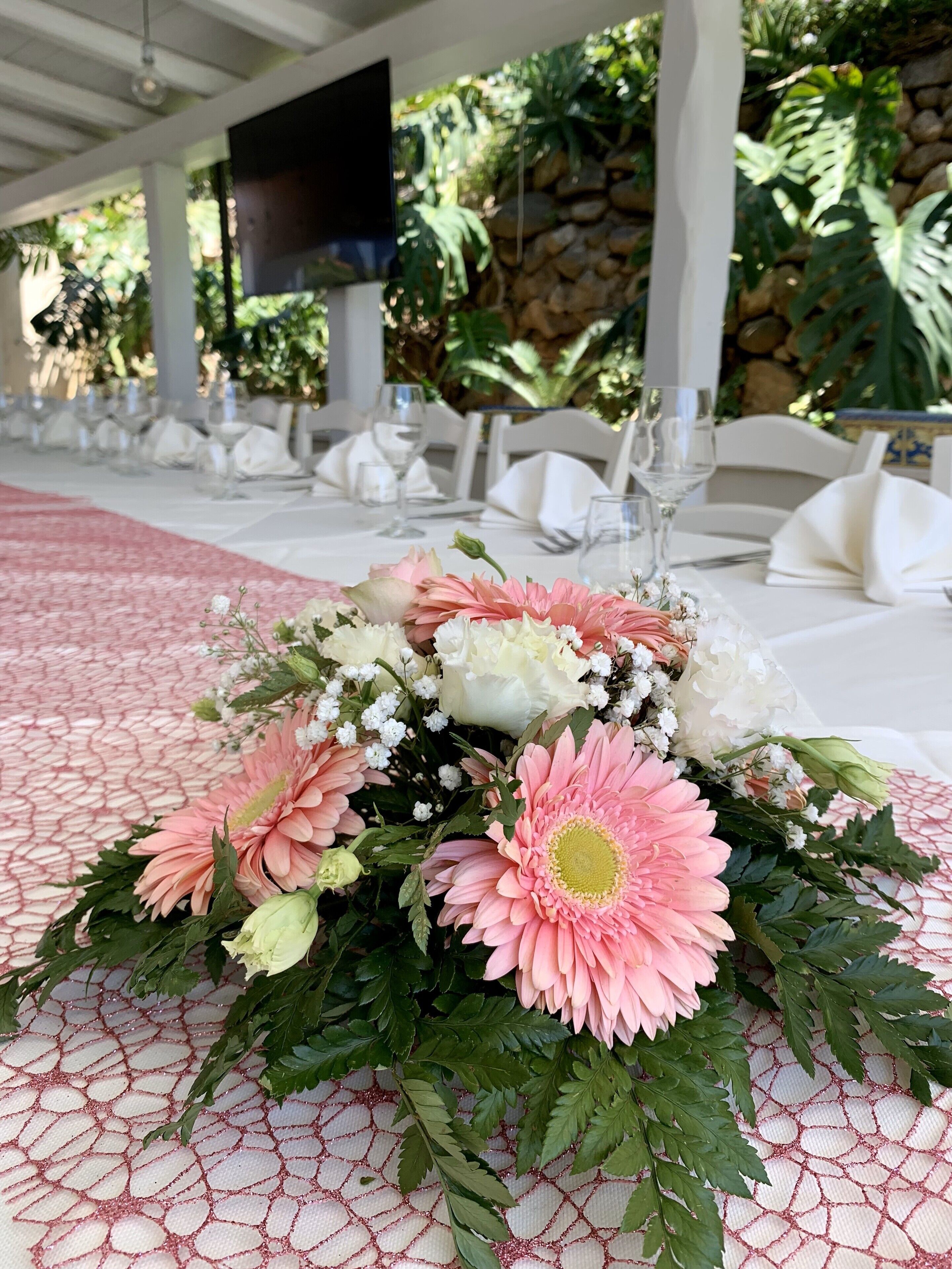 Mesa dispuesta para un evento formal, con un centro de mesa floral, mantel blanco y una pared de piedra al fondo.
