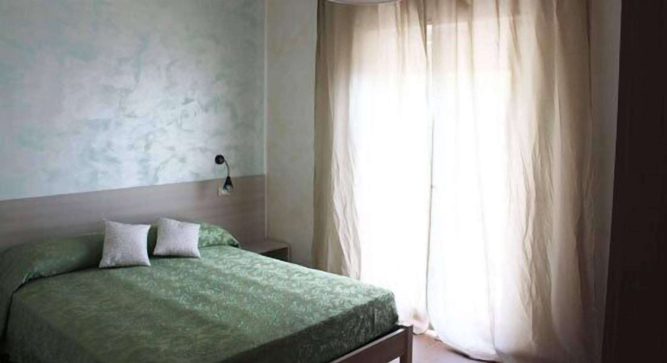 A room with a bed, a nightstand, a wall-mounted lamp, and a window with curtains.