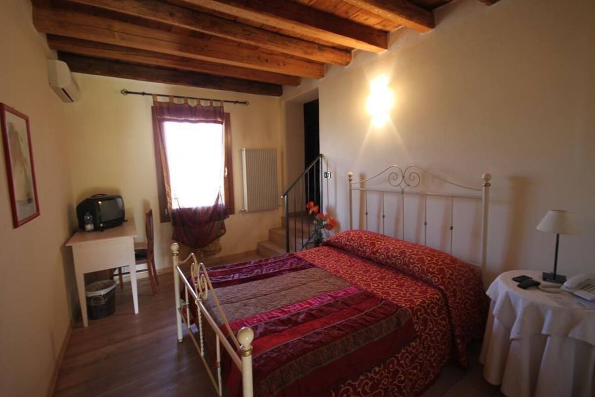 A bedroom with a wooden bed frame, a bedside table with a lamp, a small desk with a television, and a window with curtains.