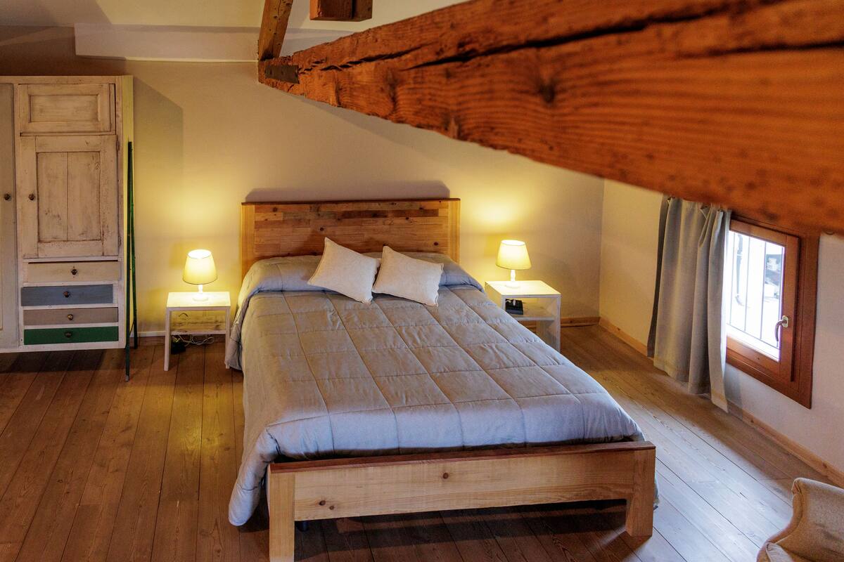 A bedroom with a wooden bed, two bedside tables with lamps, a wooden wardrobe, and a window with curtains.