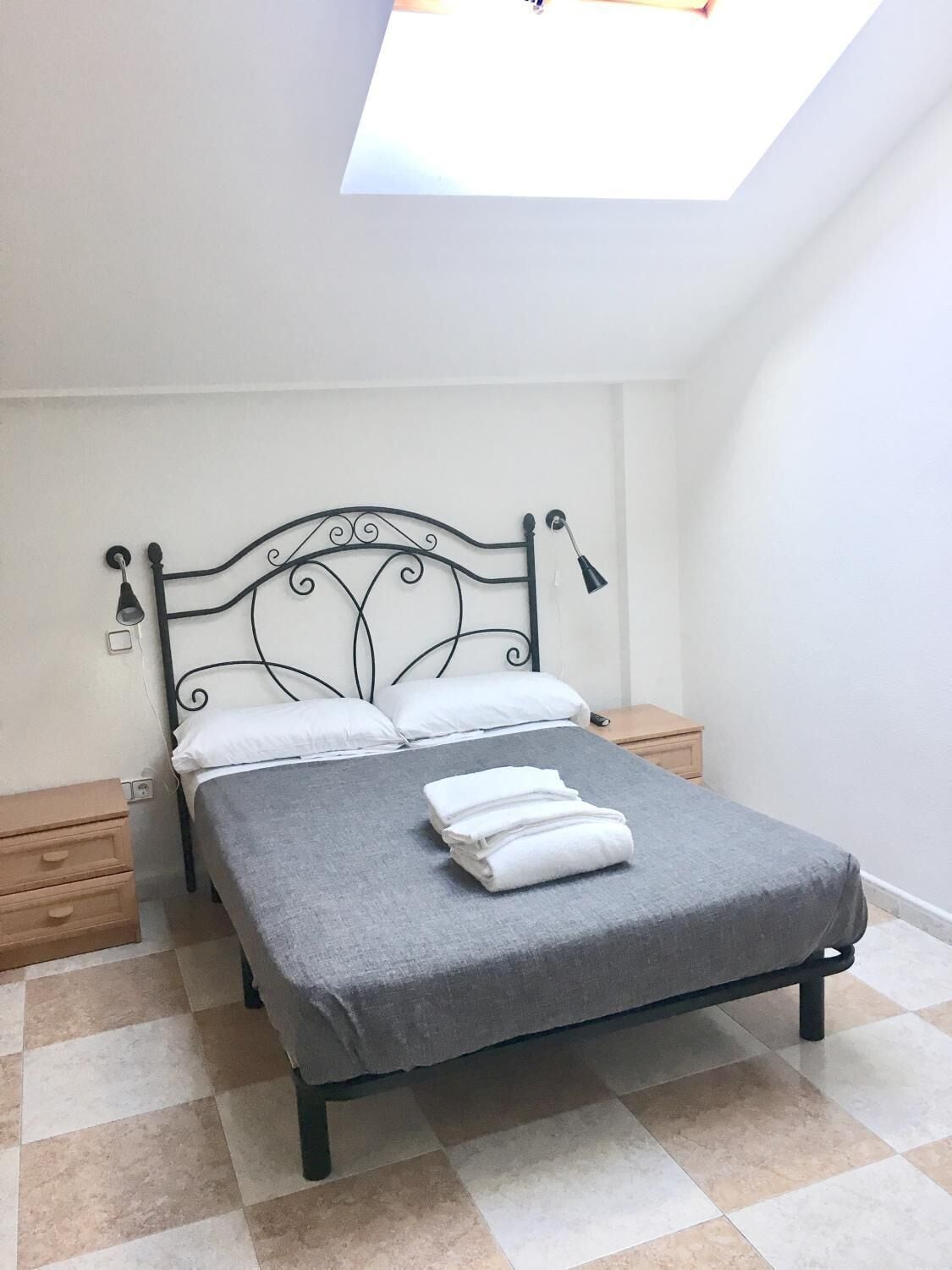 A single bed with a grey bedspread and white pillows, a small wooden nightstand, a wall-mounted lamp, and a skylight above.