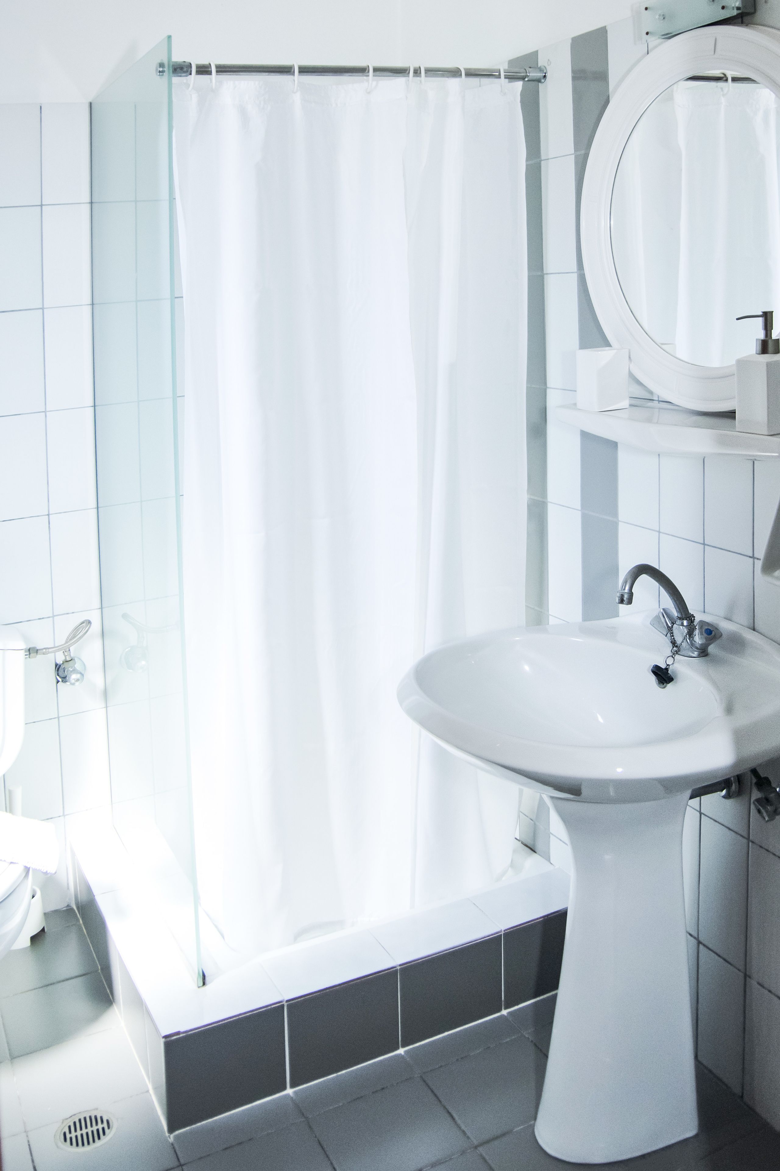 A modern bathroom with a glass shower enclosure, a pedestal sink, and a round mirror.