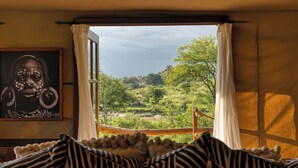 A room with a large window, a framed artwork on the wall, and a bed with striped bedding. - Elewana Serengeti Migration Camp (Serengeti)