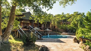 A pool area with stone flooring, a wooden deck, and a building with a covered patio. - Elewana Serengeti Migration Camp (Serengeti)