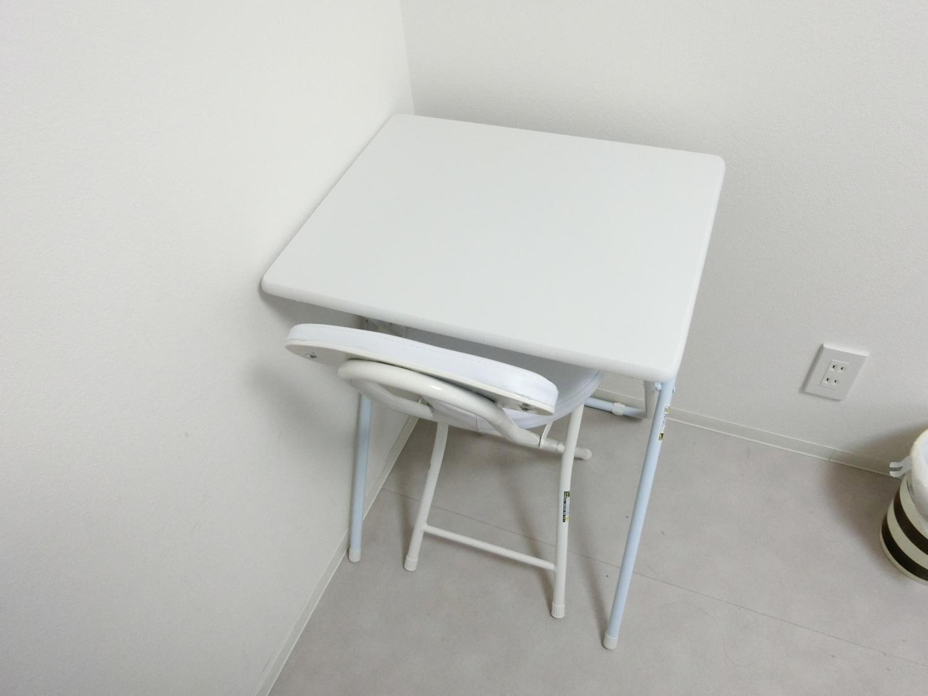 Une table blanche avec une chaise, placée dans un coin d’une pièce aux murs et au sol blancs.