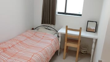 A small, clean room with a single bed, a desk, and a chair. There is a window with curtains, a radiator, and a picture frame on the desk.