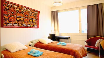 A hotel room with two beds, a desk, a chair, a large tapestry, and a window with curtains.