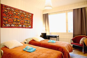 A hotel room with two beds, a desk, a chair, a large tapestry, and a window with curtains. - Wonderful Helsinki Apartment (Helsinki)