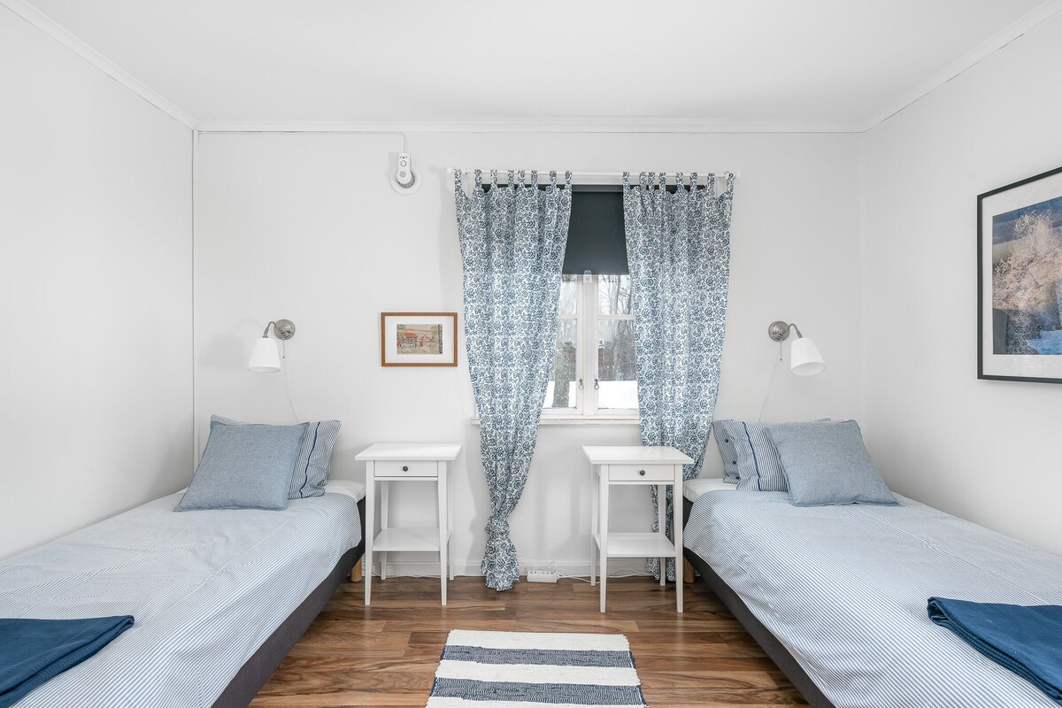 A small, clean room with two beds, a window with patterned curtains, a desk, and a rug.