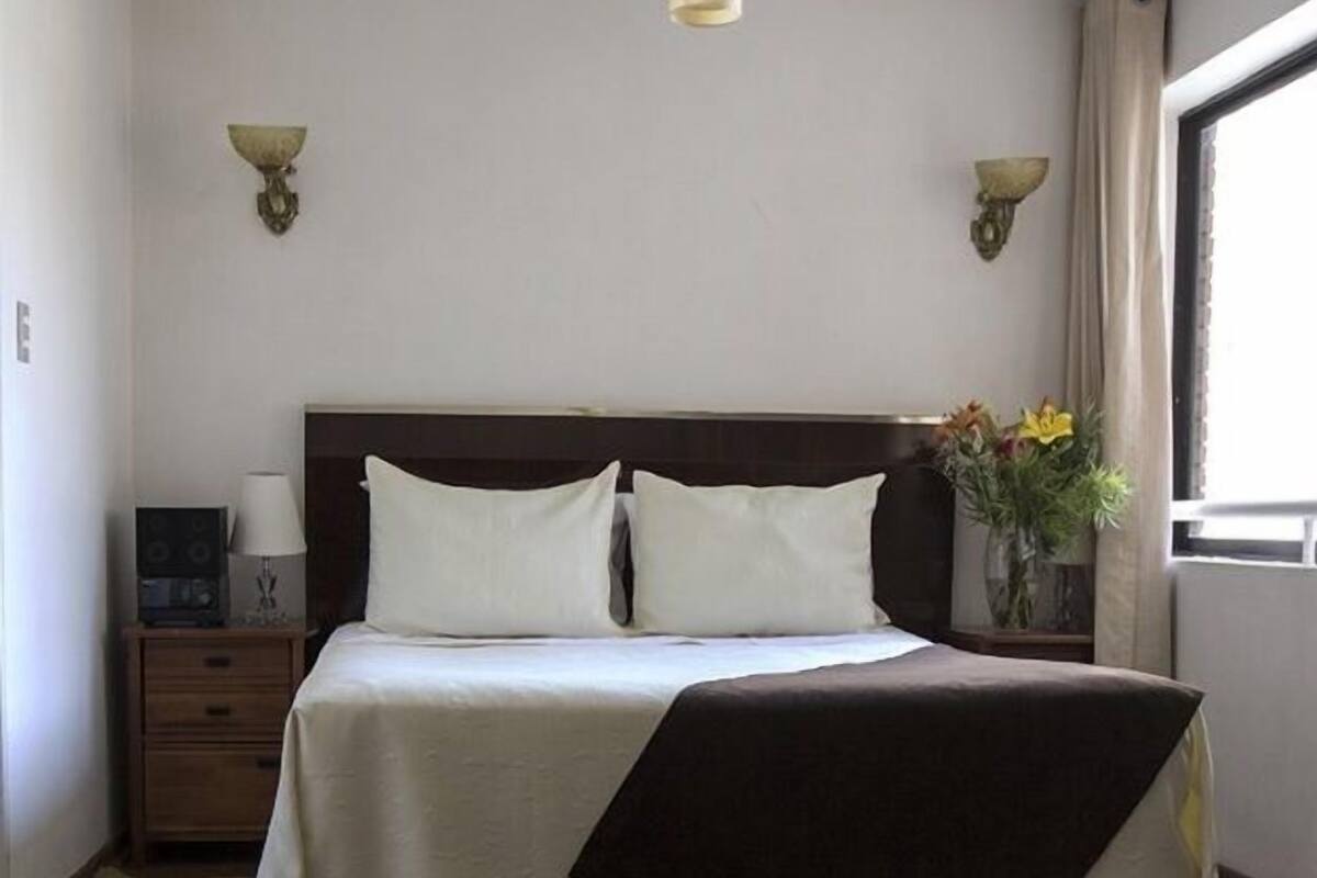 A neatly made bed with a dark headboard, two bedside lamps, and a vase with flowers on a nightstand.