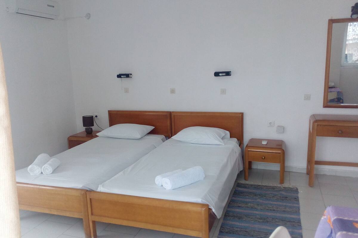 A double bed with white linens and towels, a bedside table with a lamp, a small wooden nightstand, a mirror, and a wall-mounted air conditioner.