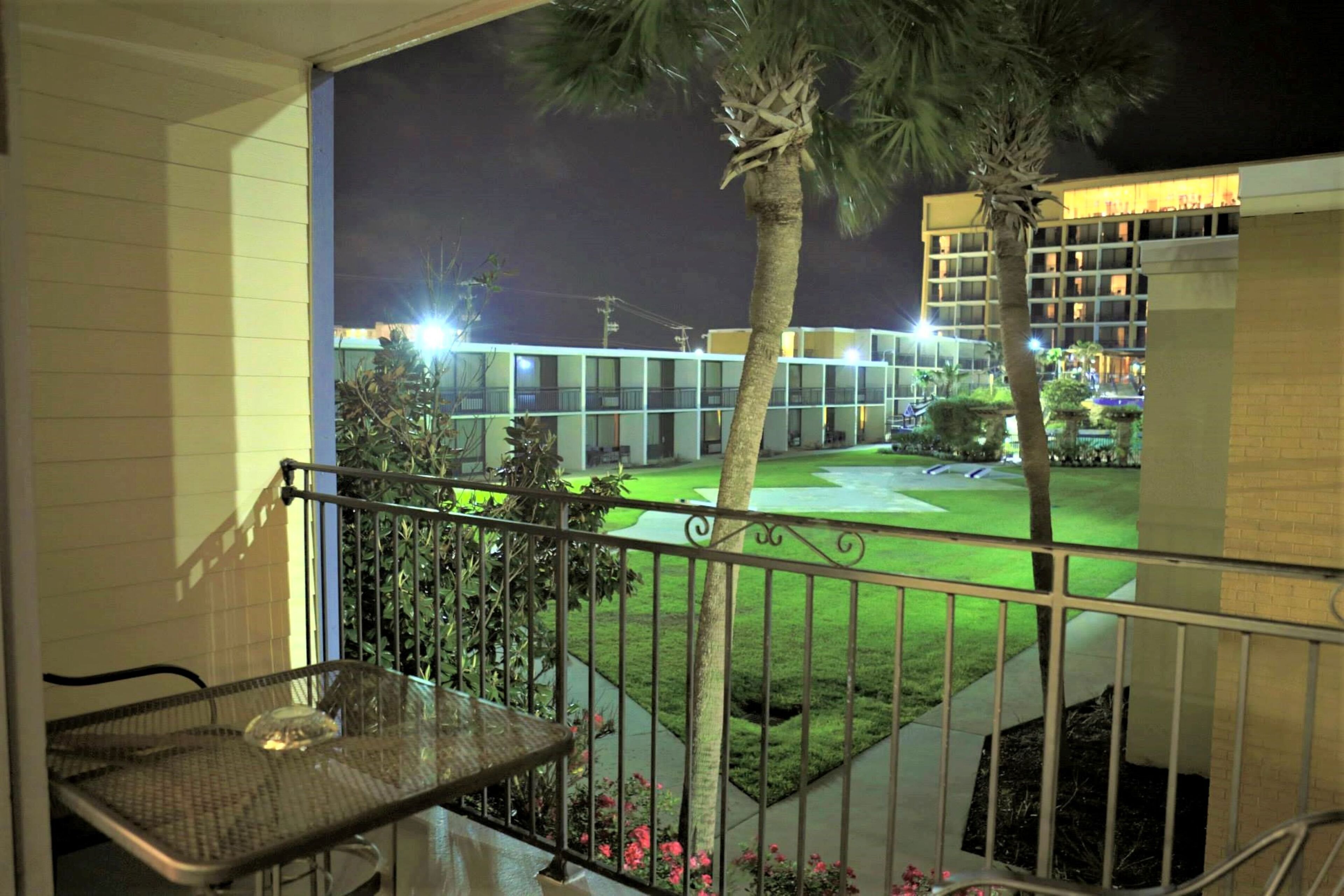 Un balcon offrant une vue sur un ensemble d’immeubles bien éclairés la nuit, avec une pelouse verdoyante et un palmier.