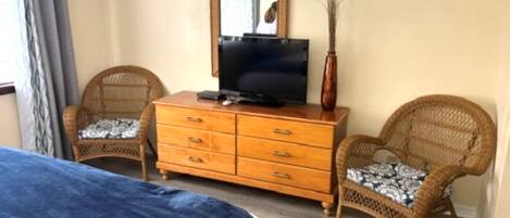 Une chambre à coucher avec un lit, deux fauteuils en osier, une commode en bois avec un téléviseur et un miroir au mur.