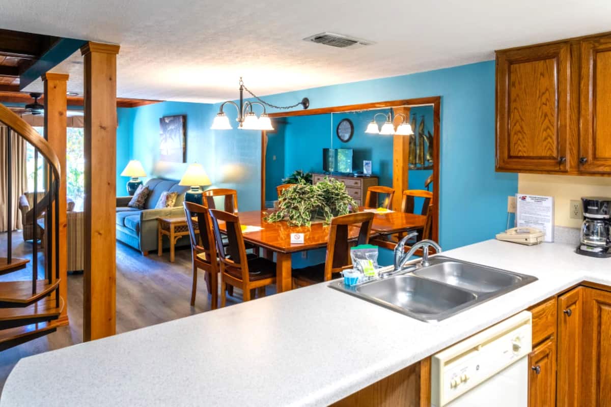 A kitchen with wooden cabinets, a dining table, chairs, a sink, and a staircase leading to another room.