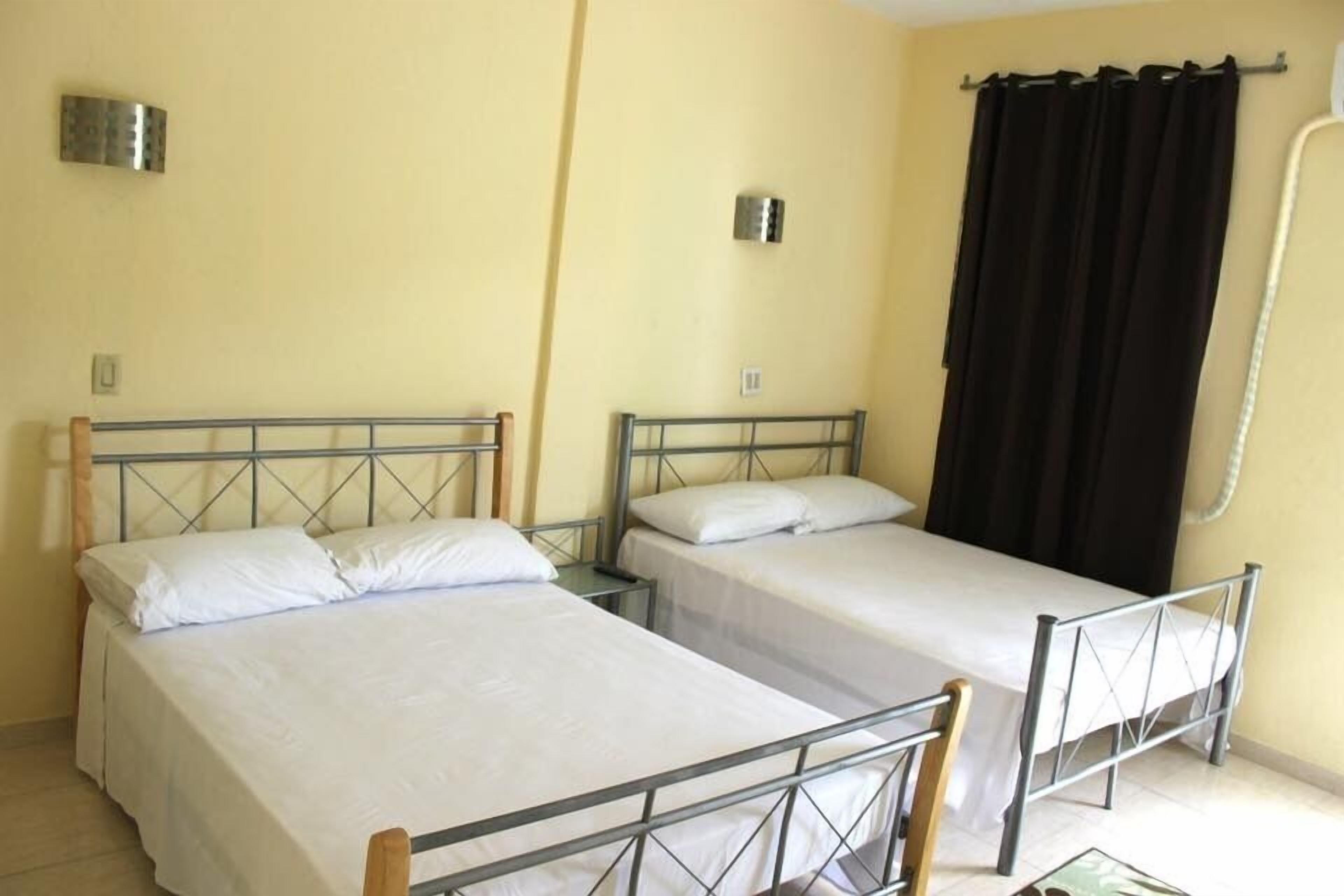 Una habitación con dos camas individuales, una ventana con cortinas negras y una lámpara fijada a la pared.