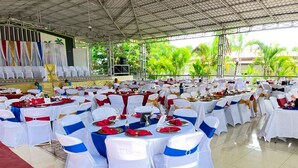 Un salón de banquetes con mesas redondas dispuestas para un evento formal, con sillas blancas y azules, manteles rojos y un escenario con fondo decorativo.