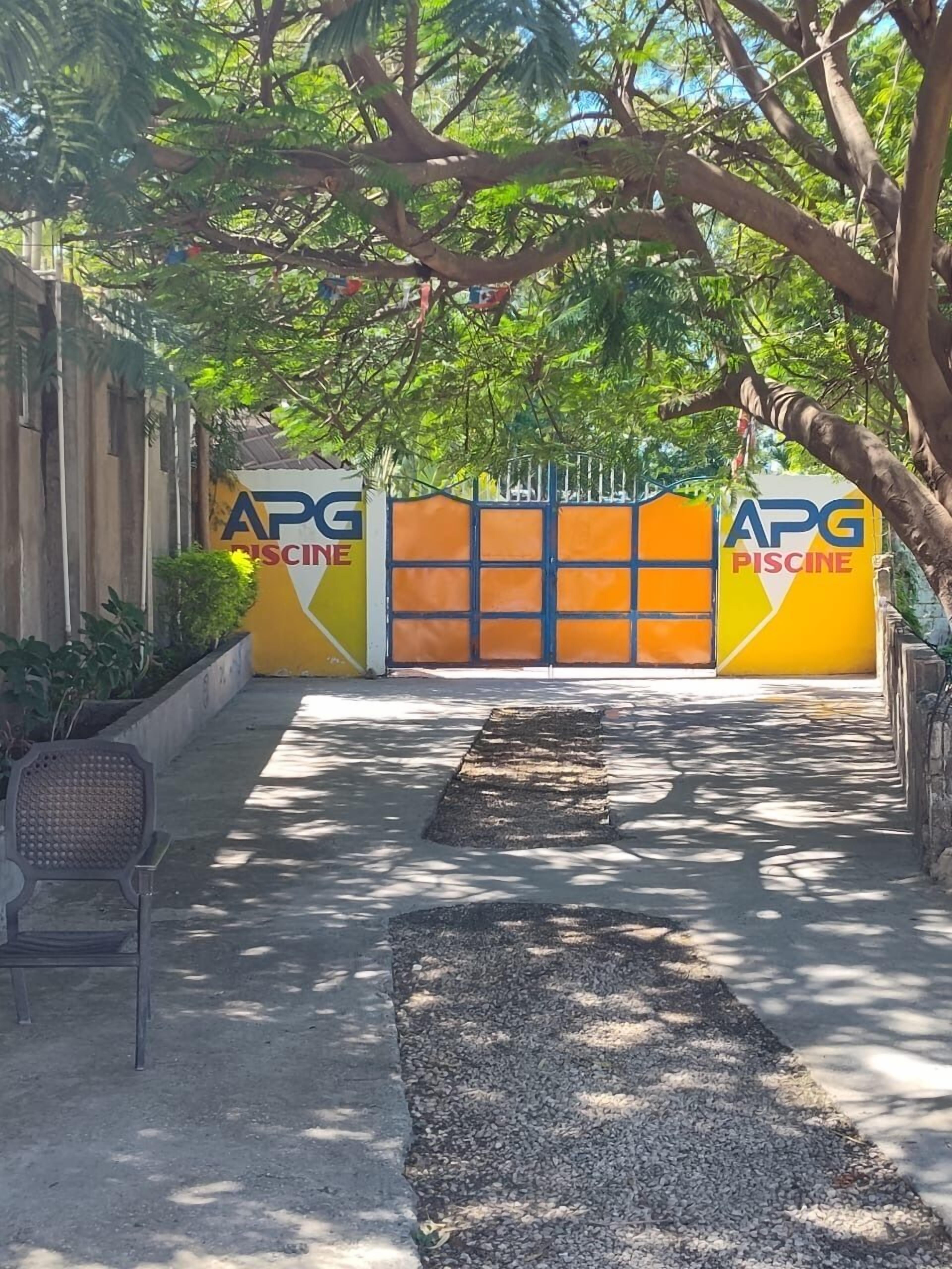 Una entrada tancada amb un mur groc i taronja, una panxa i un camí que condueix a un edifici amb la indicació 'APG PISCINE'.
