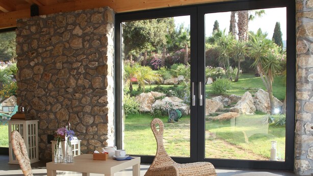 A covered patio area with wicker furniture, a stone wall, and a view of a landscaped garden. - Ulivo Blu Casa Vacanze (Castellammare del Golfo)