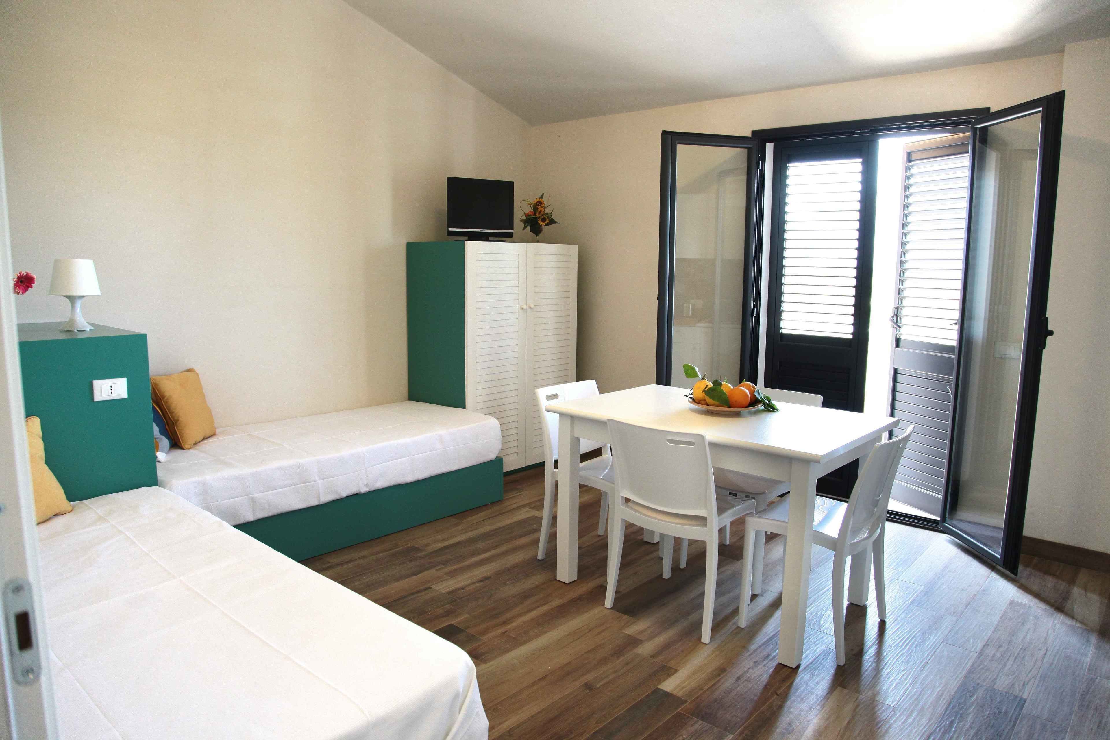 A small, well-lit room with a bed, a desk with chairs, a television on a cabinet, and a sliding glass door leading to a balcony.