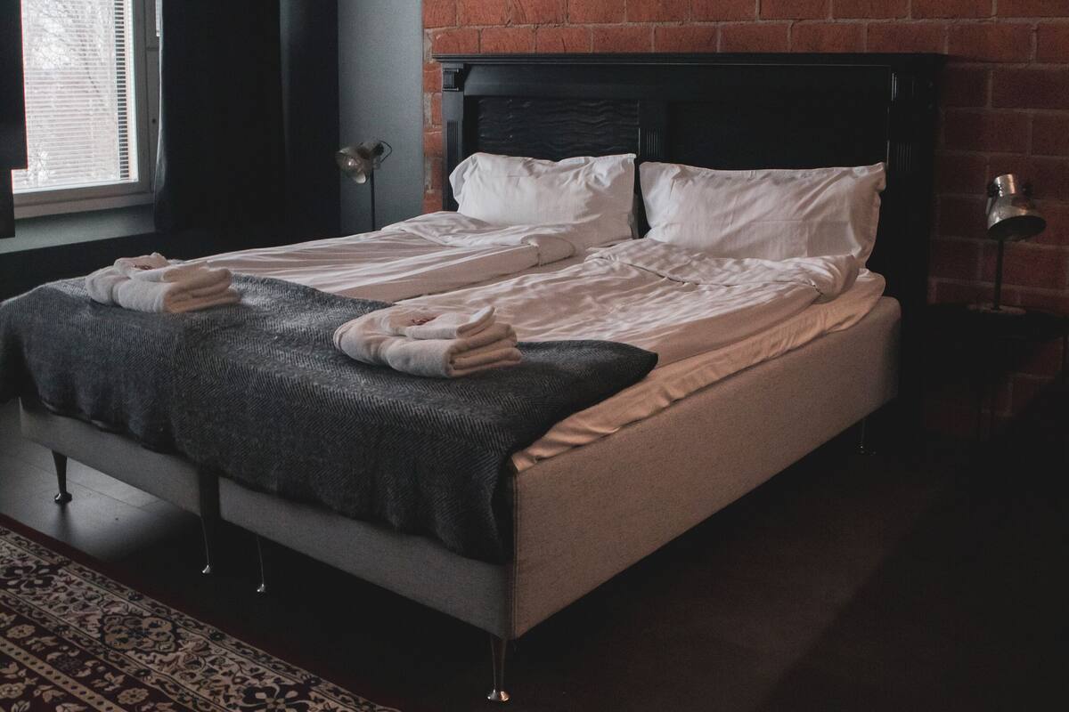 A bedroom with a brick wall, a bed with white linens, a dark rug, and a window with blinds.