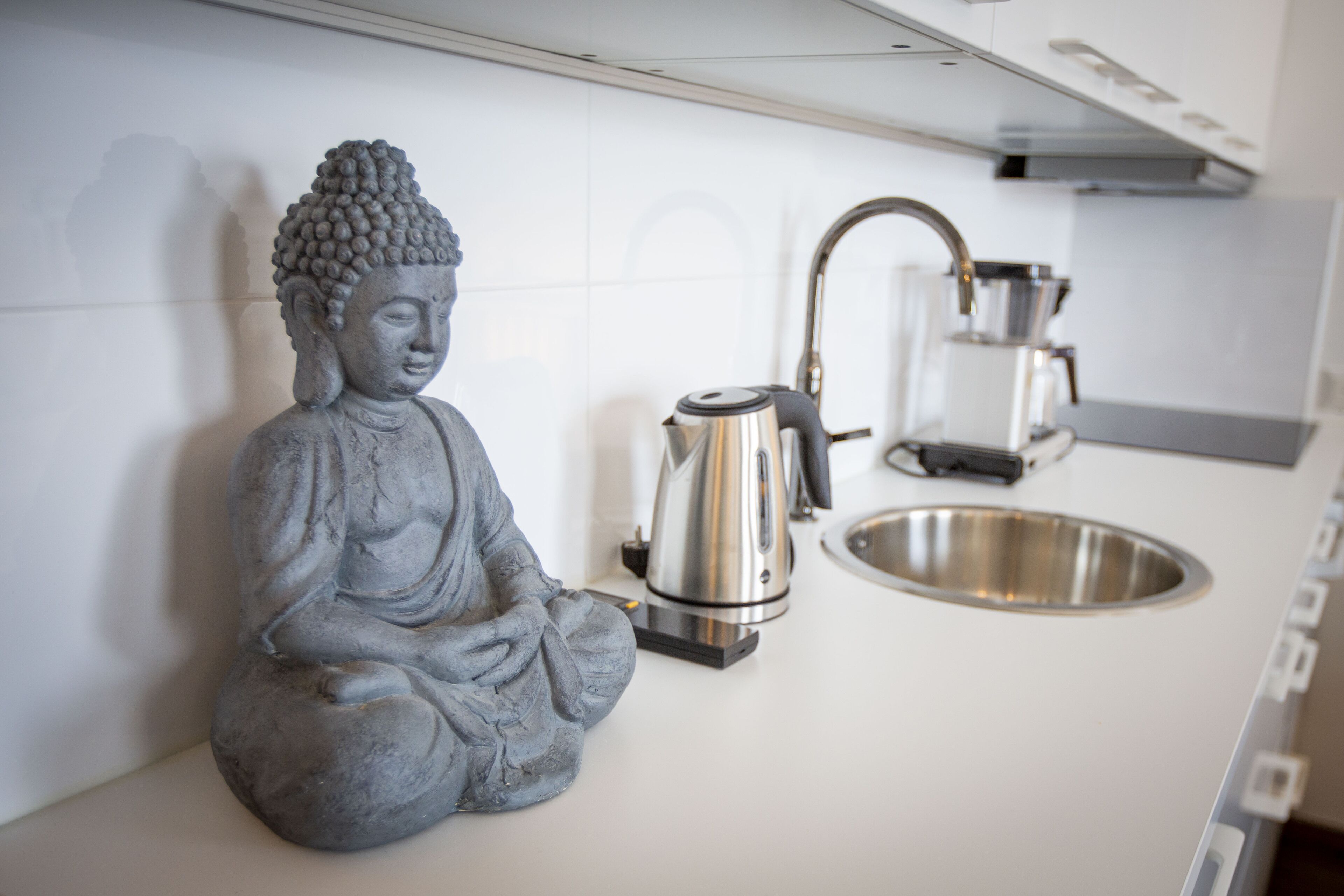 A kitchen with a Buddha statue, a kettle, and a sink.