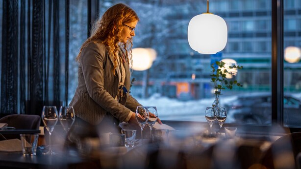 A person seated at a table with wine glasses, a vase with a plant, and a city view through large windows. - Hotel Olof (Tornio)